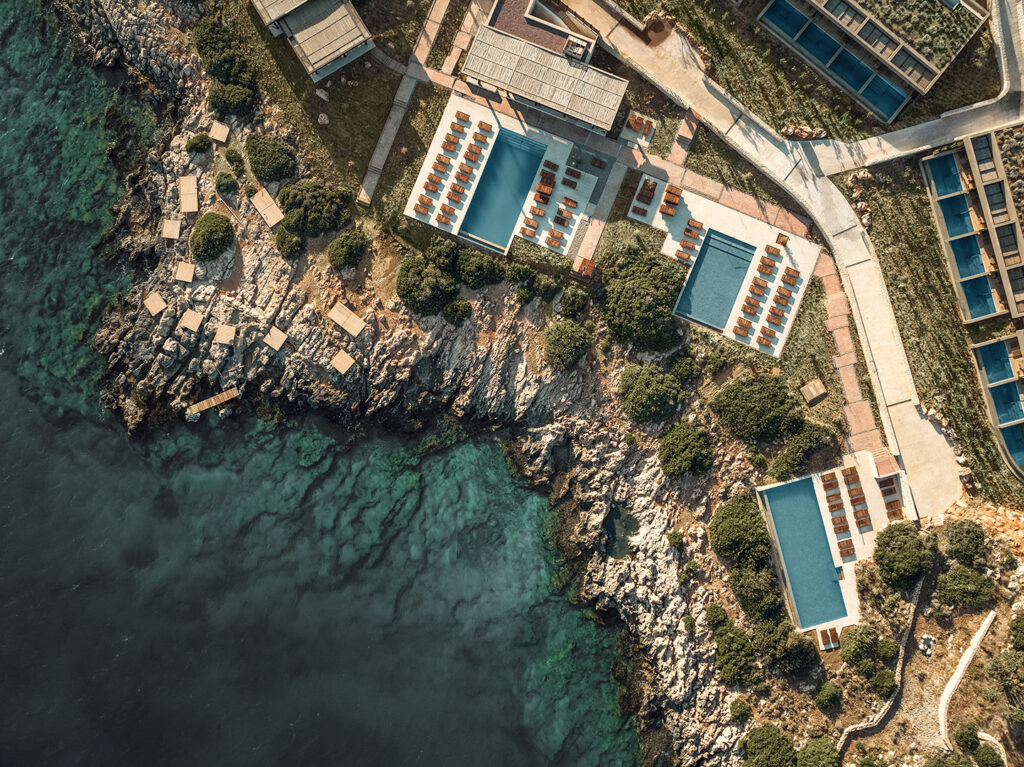 Aerial view of JW Marriott Crete Resort & Spa by Block722, showing swimming pools and terraces set into the rocky Akra Pelegri coastline above clear turquoise water.