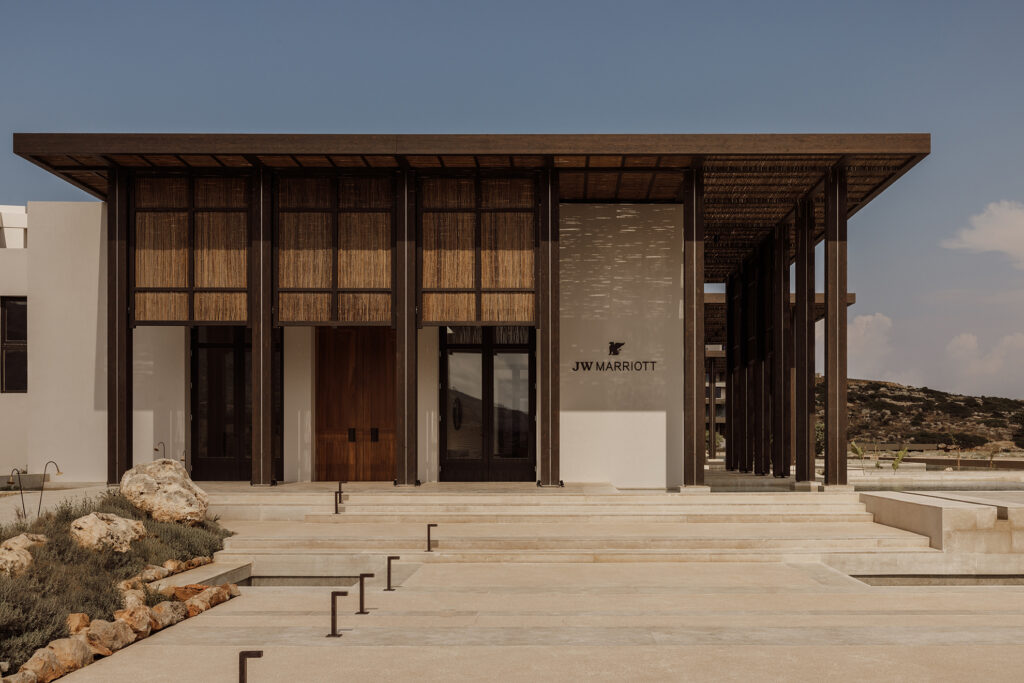 Entrance facade of JW Marriott Crete Resort & Spa by Block722, with dark timber columns, woven reed screens, and plastered walls above wide stone steps under a clear sky.