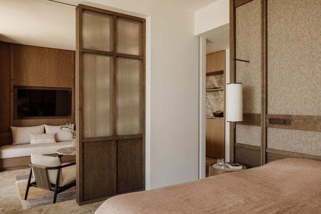 Guest room interior at JW Marriott Crete Resort & Spa by Block722, with a woven timber screen dividing a bed from a seating area of linen sofa, armchair, and wall-mounted television.