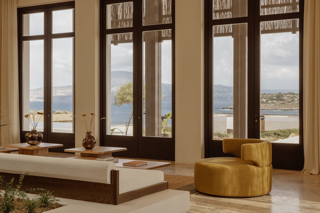 Lounge interior at JW Marriott Crete Resort & Spa by Block722, with a linen daybed, mustard armchair, and wooden tables before tall dark-framed windows opening to the sea and hills.
