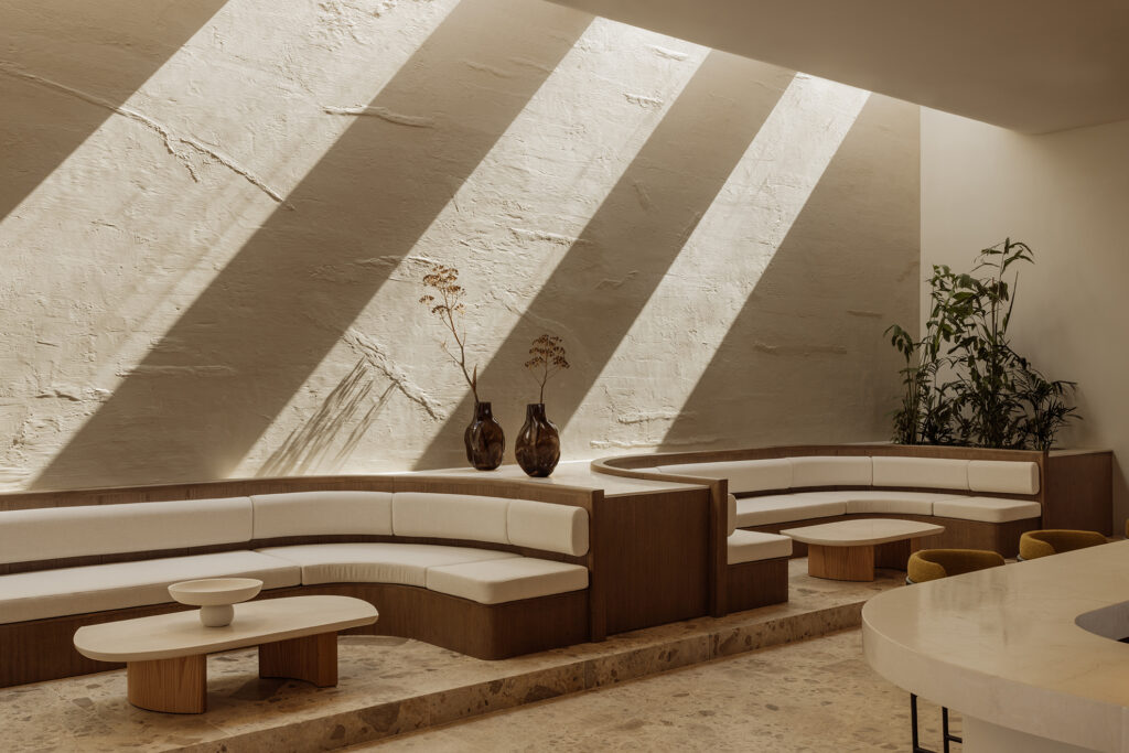 Interior lounge at JW Marriott Crete Resort & Spa by Block722, with curved built-in banquettes in cream upholstery below a textured plaster wall streaked with diagonal shafts of sunlight.