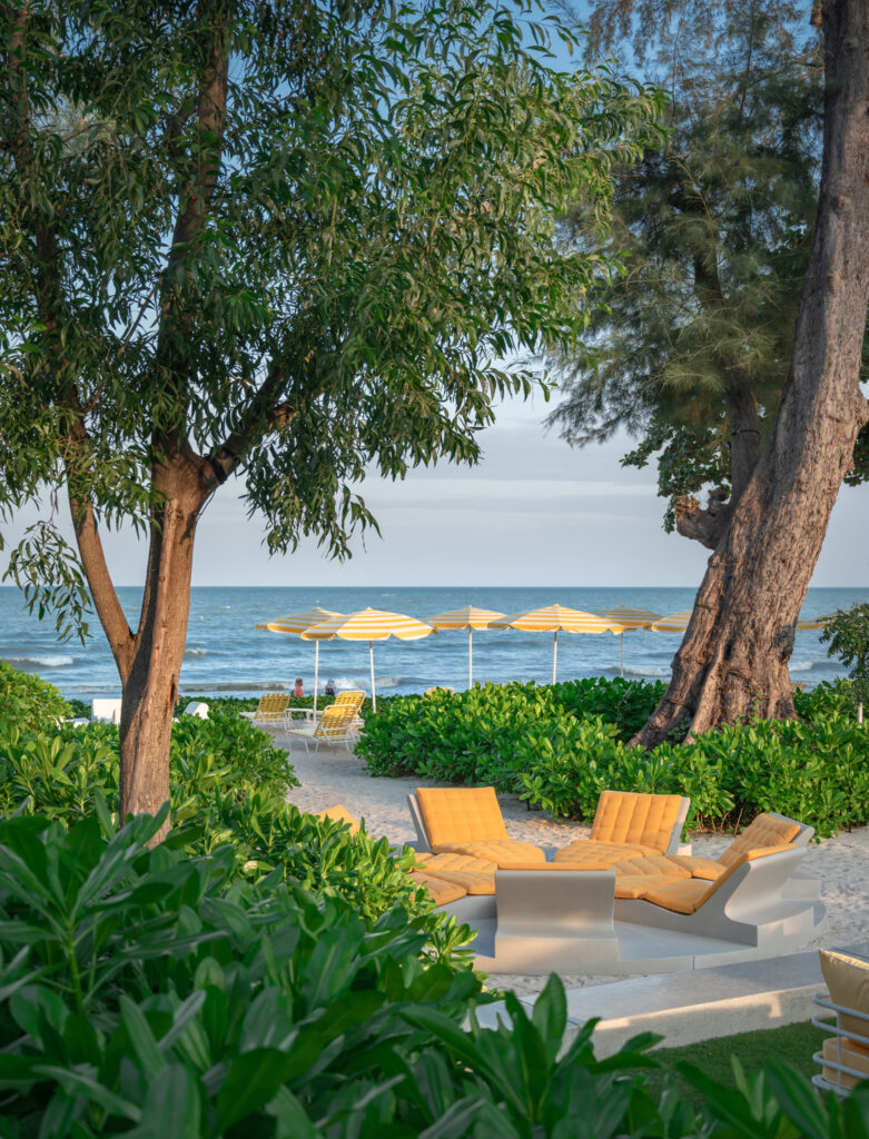 Beachfront lounge at The Standard, Hua Hin, framed by preserved mature trees, tropical landscaping by Shma, yellow umbrellas, and views of the Gulf of Thailand.
