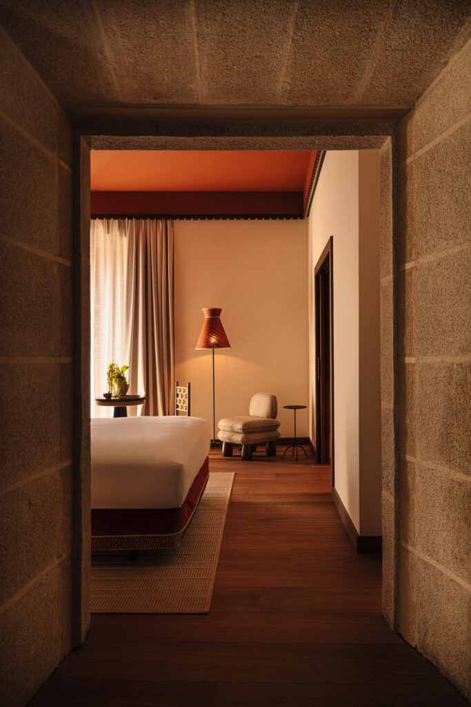 Bedroom suite at Palacio de Godoy Cáceres, framed by original granite walls, with terracotta tones and a woven floor lamp by Szymon Keller.