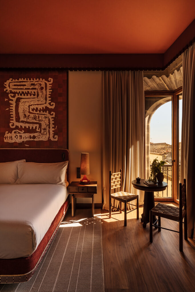 Warm-toned suite at Palacio de Godoy, Cáceres, with a tiled headboard mural, woven chairs, and an arched balcony over the rooftops.