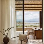Guest room interior at JW Marriott Crete Resort & Spa by Block722, with a linen armchair and stone table framing an open terrace, dining setup, and sea view toward distant mountains.