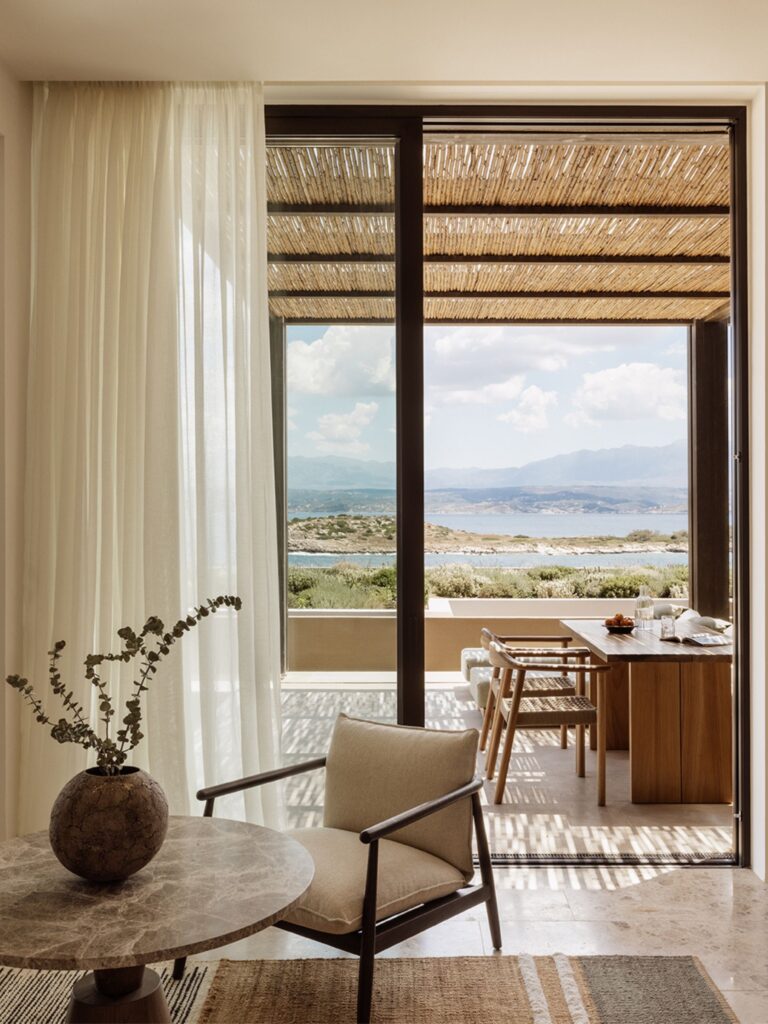 Guest room interior at JW Marriott Crete Resort & Spa by Block722, with a linen armchair and stone table framing an open terrace, dining setup, and sea view toward distant mountains.