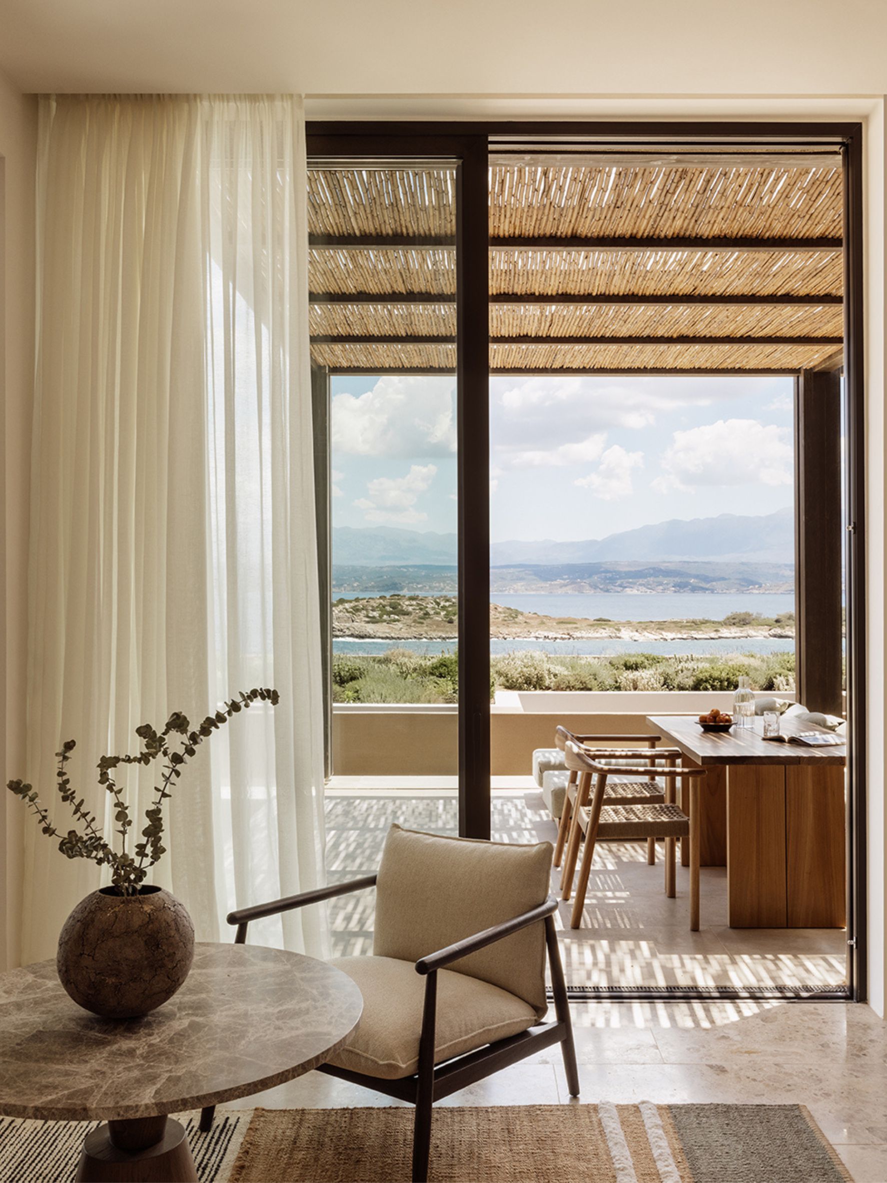 Guest room interior at JW Marriott Crete Resort & Spa by Block722, with a linen armchair and stone table framing an open terrace, dining setup, and sea view toward distant mountains.