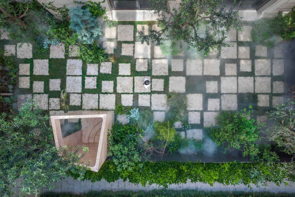 Overhead view of a visitor on grass-jointed stone paths amid mist, lush planting, and an angular travertine structure at Botanica Meditation Center.