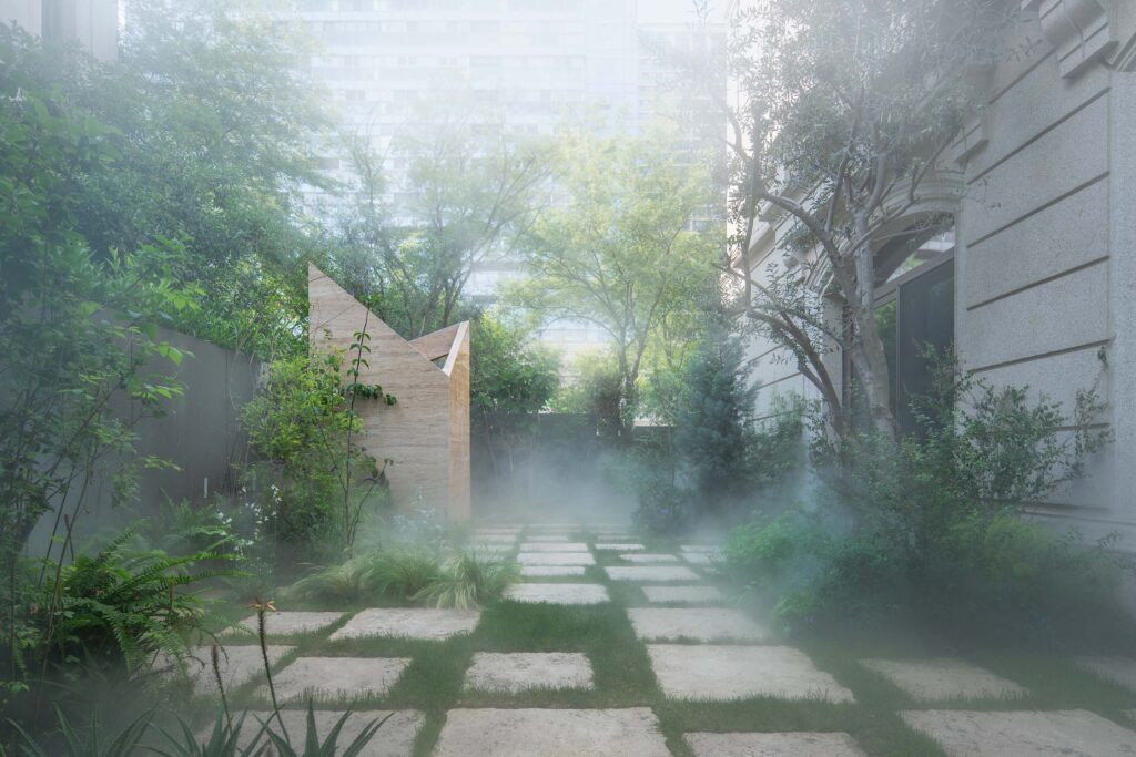 Mist drifts across a stone garden path bordered by lush plants and an angular travertine structure at Botanica Meditation Center.
