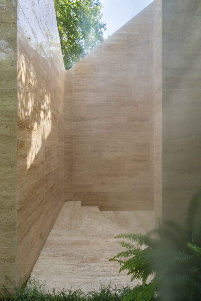 Sunlight casts leafy shadows on travertine walls and curved stone seating, with ferns in the foreground.