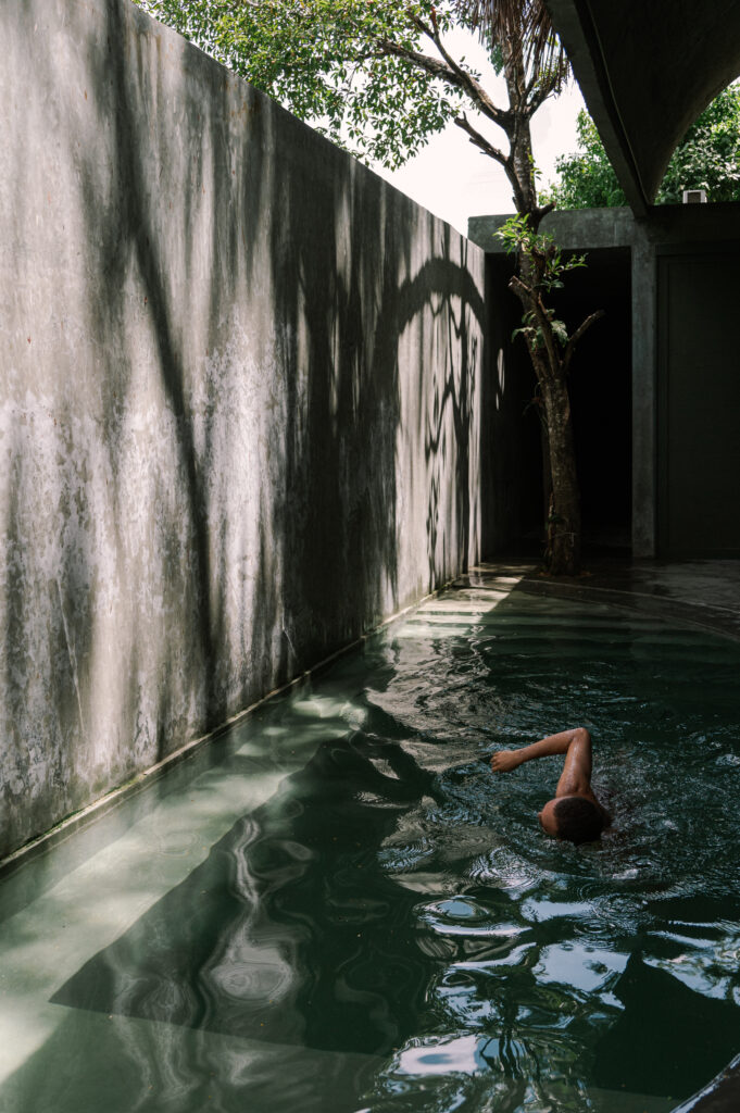 Casa Gruta in Yucatán, Mexico, featuring a person swimming in a cenote-inspired courtyard pool surrounded by textured concrete walls and tropical trees.