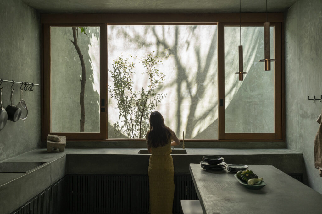 Casa Gruta in Yucatán, Mexico, featuring a minimalist concrete kitchen with wooden-framed windows overlooking a private garden.