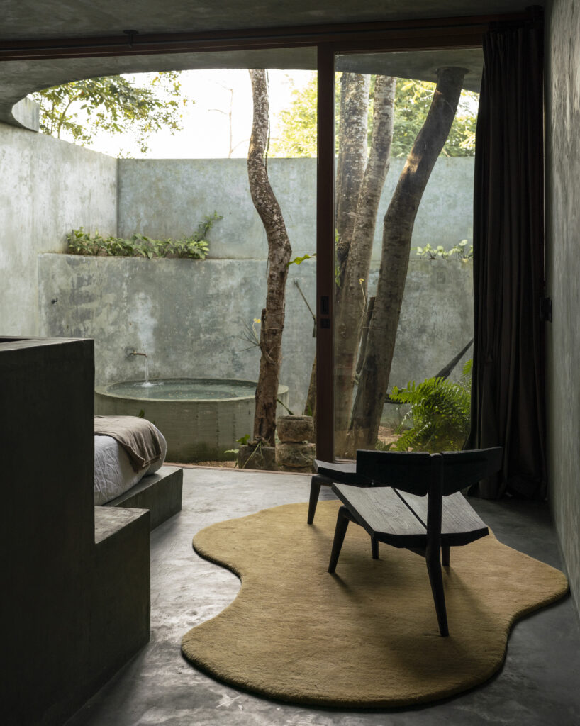 Bedroom at Casa Gruta in Yucatán, Mexico, featuring a floor-to-ceiling glass doors overlooking a private courtyard with tropical trees and a circular outdoor bathtub.