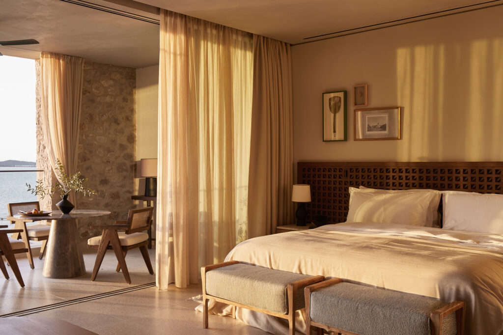 Sunlit guest room at Mandarin Oriental, Costa Navarino, Greece, with a lattice headboard and a terrace overlooking the sea at sunset hour.